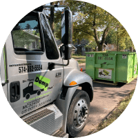 A junk removal franchise dump truck is parked beside a green dumpster on a residential street.