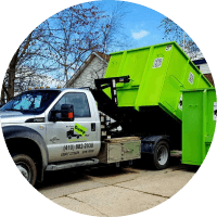 A green dumpster from a junk hauling franchise being loaded onto a white truck equipped with a hydraulic lift system.