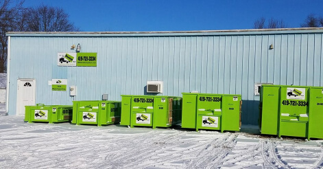 A row of green junk removal franchise dumpsters by a light blue building with snow on the ground.