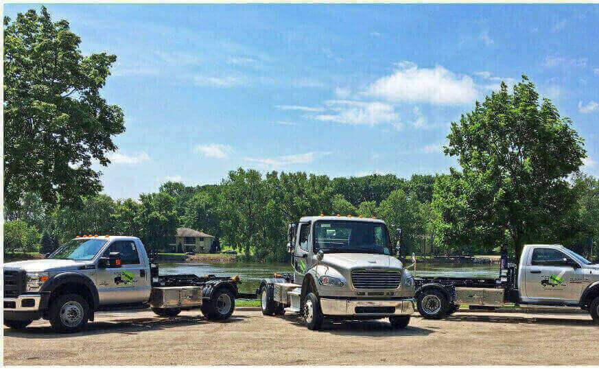 Three towing trucks parked in a row by a lake with trees, operating as part of a junk hauling franchise on a sunny day.