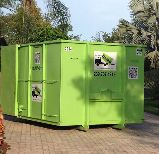 Green dumpster from a junk hauling franchise, situated outdoors near vegetation.