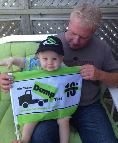 A baby wearing a cap sits on a green chair with an adult, both holding a flag of a junk hauling franchise print and text.