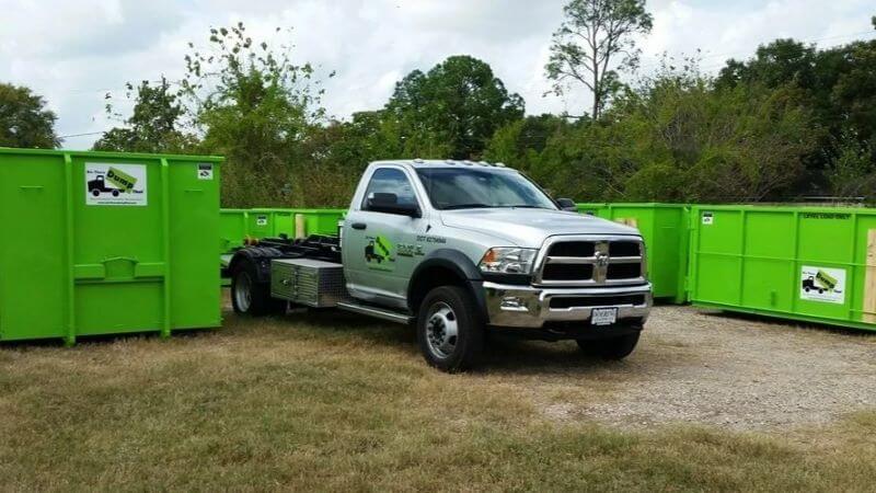 A white pickup truck is towing a trailer with two green dumpsters from a dumpster rental franchise against trees and grass.