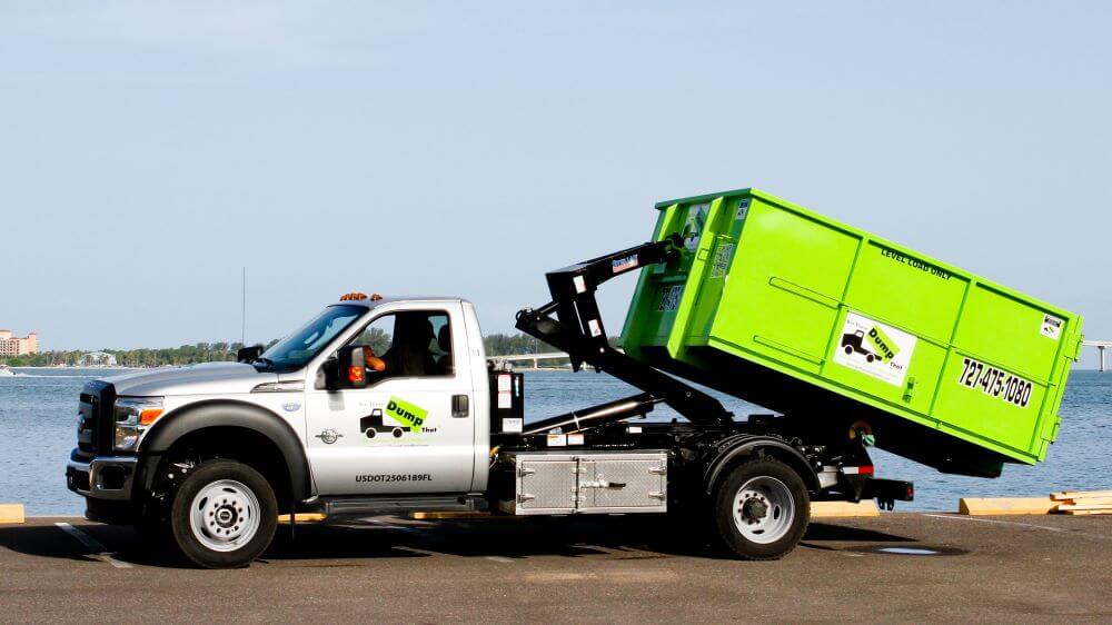 A green waste management dumpster rental franchise being loaded onto a white truck by the waterfront.