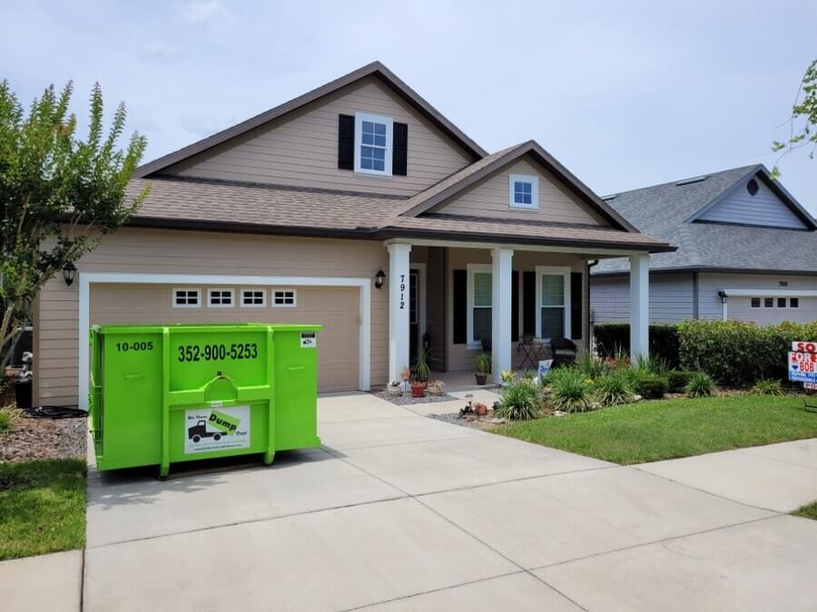 A beige single-story house with a lawn and a large green dumpster franchise in the driveway, under a clear blue sky.