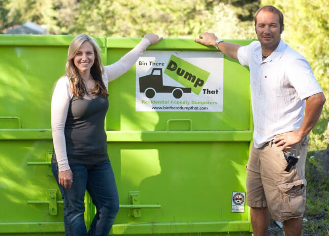 Two people standing beside a green dumpster with a "bin there dump that" logo from a dumpster rental franchise.