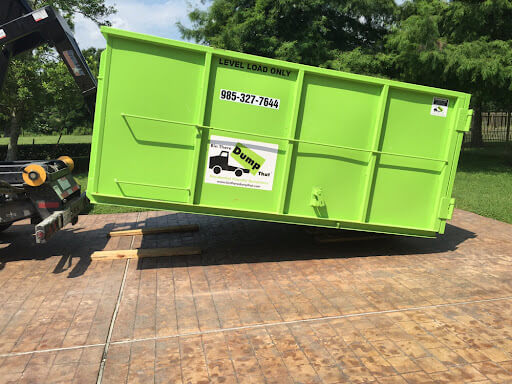 A green dumpster from a low-cost dumpster rental franchise, with a "level load only" sign and a phone number, is parked on a wooden platform, with a black tow truck visible to the side