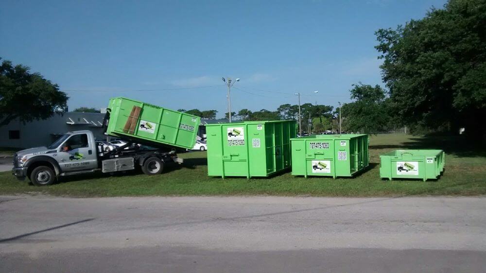 A truck is unloading a green dumpster onto a grassy area next to several other green dumpsters, illustrating the seamless operations of owning a dumpster business. Trees and a building are in the background, framing this efficient scene.