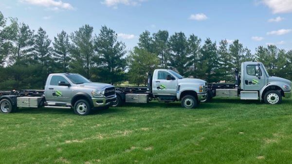 Three silver tow trucks are parked on a grassy field with a line of trees in the background.