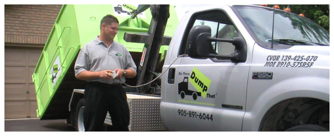 A man stands next to a white truck with a green dumpster, holding a remote control. The truck's doors display company information and a logo that reads 